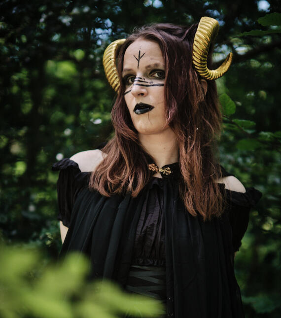 Portrait von Lilischote in einem schwarzen Waldgeist Kostüm. Sie trägt blasses paganisches Makeup mit Runen und goldene Hörner.